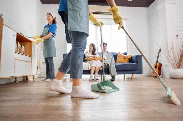 a couple sitting on a couch while two cleaners are cleaning the living room
